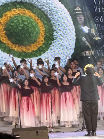 This image captures an indoor stage performance, likely a choral or musical event, in Guta, China. The scene features a group of young performers arranged in rows on a stage, backed by a large digital screen displaying a vivid, abstract circular pattern resembling a flower or target, with concentric rings of green, yellow, and blue hues.
The performers consist of both male and female individuals. The women wear black tops paired with elegant, layered skirts that exhibit a white-to-pink/red ombre effect. The male performers are dressed in lighter-colored shirts (possibly white or light blue) accessorized with bow ties. All performers are holding illuminated, star-shaped objects, extending them forward as a synchronized visual element of the act.
At the forefront, a conductor or leader, dressed in a dark suit, stands with their back to the camera, facing the performers and seemingly directing the ensemble. Microphones on stands are visible near the performers, confirming a vocal component to the performance. Stage equipment, including a dark speaker, is situated on the right side of the stage.
The background screen also projects images of other figures, including what appears to be a person in a military-style uniform. Fragments of text are visible on the screen, including repeated "Vit" and "Vict" possibly indicating "Victory," along with the Greek letter Theta (Θ). The setting suggests an evening event, with artificial lighting illuminating the stage and performers. The participants appear focused and engaged in their performance.