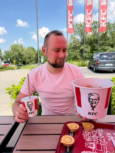 A man with a beard and short dark hair, wearing a light pink t-shirt, is captured enjoying a meal outdoors at a KFC restaurant. He is seated at a wooden table, holding a large white KFC bucket containing small pieces of fried chicken in one hand and a smaller white and red KFC cup with a straw in the other.
On the table next to him, a red plastic tray holds three small cups of "KFC Kentucky Gold" sauce. The tray also features Polish text, "GRAMY DO JEDNEGO KUBELKA" (WE PLAY FOR ONE BUCKET), and "OFIJAL MISTR CE 3x3," likely promoting a 3x3 event.
The background reveals a bright blue sky with white clouds, and lush green trees. Several red flags displaying the "KFC" logo in white are mounted on poles, reinforcing the brand presence. A dark-colored car is parked in the distance. The scene is set on a clear, sunny day, indicating daytime. The man appears relaxed, possibly enjoying the outdoor dining experience at this KFC location, identified as being in Ustroń, Poland.