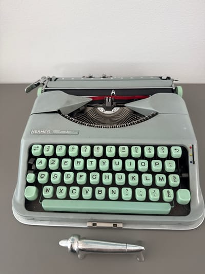 The image displays a vintage Hermes Baby typewriter, pale green in color, positioned on a grey surface. The typewriter features a standard QWERTY layout with light green keys, and the "HERMES Baby" logo is visible on the front. Below the typewriter, a shiny metal object, possibly a pen or a decorative item, rests on the surface. The background is a plain white wall. The scene is set in Drumettaz-Clarafond, France. No people are present in the image, and there are no discernible actions or interactions taking place. The environment appears to be indoors, in a well-lit space.