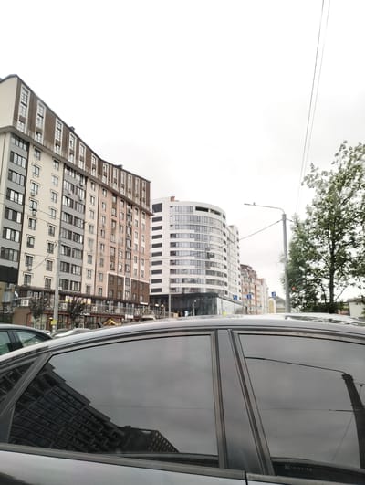 The image presents an urban street scene in Ivano-Frankivsk, Ukraine, viewed from inside or beside a dark-colored vehicle on an overcast day. The upper part of the car, including its roof and dark tinted windows, occupies the bottom portion of the frame. The windows reflect the muted sky and buildings opposite the viewer.

The background is filled with several multi-story buildings. On the left, a large, modern building with a curved facade displays a combination of light-colored, grey, and brown sections, featuring numerous rectangular windows and balconies. Below this building, a commercial storefront is visible, with the text "КАЛЬВ" (KALV) appearing on its signage. Adjacent to this, a prominent, modern white building with a rounded, cylindrical design stands out, characterized by its grid of dark windows. Further down the street, additional buildings with varying architectural styles can be seen.

To the right, a street light pole with overhead wires extends into the frame, partially obscuring a tree with green foliage. The sky is uniformly bright and overcast, suggesting daytime without direct sunlight. There is no explicit activity taking place, but the presence of multiple buildings, vehicles, and infrastructure conveys a bustling city environment. Faint text, "NC 18-XX-YB", is visible on the bottom right car window.