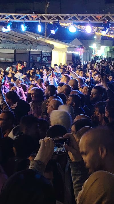 The image captures a large crowd of people attending an outdoor event at night in Ash-Shaghour Municipality, Syria. The scene is brightly illuminated by artificial lights, including several prominent blue stage lights mounted on a metal truss structure across the top of the frame. Decorative string lights are also visible further in the background, adding to the festive atmosphere.

The densely packed crowd consists mainly of adults, with individuals facing generally towards the left side of the frame, suggesting a focal point such as a stage or performance area. Many attendees are actively engaged, holding up smartphones to record videos or take photographs of the event. White tent-like structures are visible in the background, hinting at stalls or a market setup typical of a festival or fair.

The time of day is clearly night, as indicated by the darkness of the surroundings and the heavy reliance on artificial light sources. The mood appears to be one of collective engagement and excitement.

A notable detail is the partially visible Arabic text on a sign in the upper right background. Although somewhat blurry, it can be deciphered as "مهرجان مدينة الشعور / البلدية الدورة..." which translates to "Ash-Shaghour City/Municipality Festival, Session...". This text explicitly confirms the event's location and nature as a municipal festival in Ash-Shaghour.