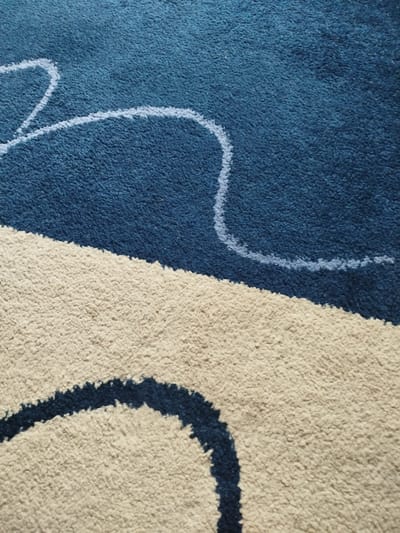 The image is a close-up, overhead shot of a carpet. The carpet features a design with two distinct color blocks. The top portion is a deep blue, and the bottom portion is a light beige. A thin, wavy white line runs across the blue section, and a thicker, wavy dark blue line cuts across the beige section. The texture of the carpet appears to be thick and shaggy. There are no people, objects, or actions visible in the image, and no discernible environmental context is provided to indicate a location in Vynnyky, Ukraine.