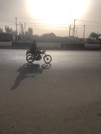 A motorcyclist performs a wheelie on a paved road. The bike's front wheel is high in the air, and smoke rises from the rear tire. A long shadow stretches across the asphalt, cast by the rider and the motorcycle. Power lines and trees line the side of the road under a hazy sky.
