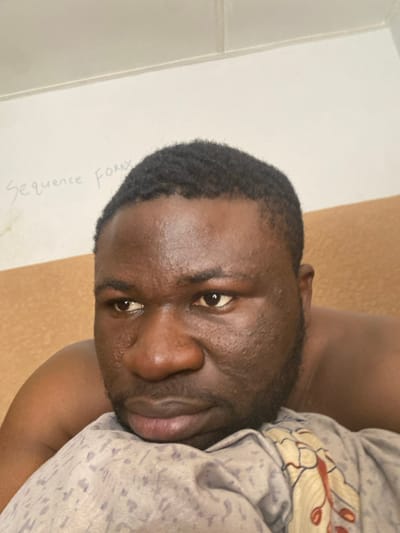 An adult male with dark skin, short dark wavy hair, and a short beard is depicted in an indoor setting within Udu, Nigeria. He is positioned with his head or upper body resting on a light grey patterned fabric, possibly a pillow or blanket, which features a faint floral or leaf design in muted colors. The man's bare right shoulder is visible, suggesting he is shirtless or partially clothed. He looks towards the left side of the frame with a neutral or contemplative expression. In the upper left background, a light-colored wall is visible, on which the handwritten text "Sequence Forex" is faintly discernible. A solid light brown or beige wall surface is present behind and to the right of the man.