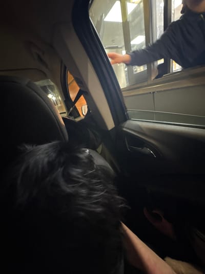 Inside a car at a drive-through, a person reaches toward a window to receive an item. Another person sits in the passenger seat. The scene is darkly lit, but light shines from outside, illuminating the interaction at the window. The car interior is visible, including the dashboard, seats, and headrest.
