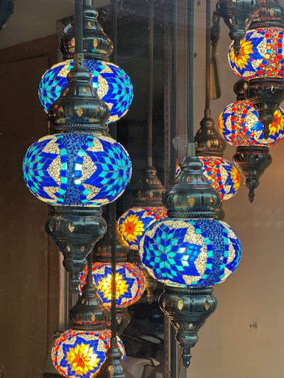 The image shows a row of decorative mosaic lamps reflected in a dark, glossy surface. The lamps alternate between blue and orange/red color schemes, each comprised of many small, patterned tiles. The lamp fixtures appear to be made of dark metal. The scene is presumably indoors, given the controlled lighting and the reflective surface. The location in the city of Wisła, Poland is not visually evident in the image, so information about the location is not possible to ascertain. No people, actions, or interactions are visible.