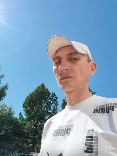 This is a close-up, low-angle selfie of a man wearing a white baseball cap and a white t-shirt with a repeating black and grey graphic print. He has short brown hair, brown eyes, and a thin mustache. The man is looking directly at the camera with a neutral expression.
The background is a clear, bright blue sky with some lens flare from the sun in the upper left corner. Below the sky, there are dark green evergreen trees, suggesting an outdoor setting, likely in a natural or park-like environment. The presence of trees and the bright daylight indicate it is daytime, likely sunny.
The visible text on the t-shirt appears to be a stylized graphic that repeats multiple times. While the exact wording is difficult to decipher completely, it seems to be an abstract or branded word.
The image was taken in Bor, Russia, based on the provided location context. The bright sun and blue sky suggest pleasant weather conditions. The overall mood of the image is casual and straightforward.