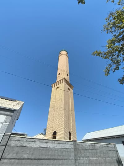 A tall, slender minaret, constructed from light yellow bricks, dominates the center of the image, reaching towards a clear blue sky. The minaret has a ribbed, green dome at its apex and features several arched openings and decorative brickwork along its height. In the foreground, a grey concrete wall runs horizontally across the bottom of the frame, partially obscuring the base of the minaret. To the left of the minaret, the corner of a building with a white, corrugated roof is visible, juxtaposed with a section of a grey and white patterned wall. To the right of the minaret, another building with a white, sloped roof can be seen. Several dark electrical wires stretch across the blue sky, traversing the scene from left to right. A cluster of green leaves from a tree branch hangs from the upper right corner of the image. The overall scene depicts an urban environment, likely within the city of Babakhodja, Uzbekistan, with a prominent religious structure.