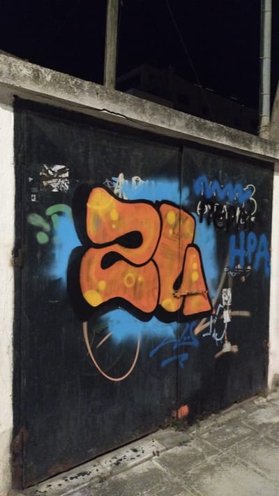 This is a nighttime, eye-level, medium shot of graffiti on a large, dark metal garage door. The graffiti features bold, orange letters with yellow highlights and black outlines, superimposed on a bright blue spray-painted background. To the right of the main graffiti, there are smaller, blue and white spray-painted tags. The garage door is set within a rough, concrete structure that extends to the left and right edges of the frame, topped by a cracked, weathered concrete lintel. Above the lintel, the dark night sky is visible, with the silhouette of a building in the distance. The ground in front of the garage door is paved with concrete tiles. The image was taken in the city of Perea, Greece.