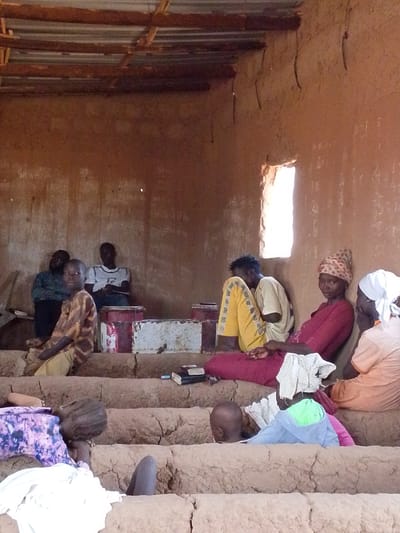 The image shows a group of people inside a structure in Mulke Barikin Sayejo, Nigeria. The building appears to be constructed of mud or clay. The roof is made of corrugated metal sheets. There is a small window providing light. A group of adults are seated, some wearing head coverings. A few children are seated on the lower level. There are books and various containers present. Some people are talking to each other.