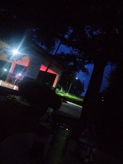 The image depicts a scene at dusk or night in Mangu Town, Nigeria. The foreground is dark and slightly out of focus, with a partially visible plastic bottle and a cylindrical object that could be a thermos or a bottle. In the mid-ground, a building or structure with white walls and a covered porch is illuminated by lights. Red light is visible inside one of the rooms of the structure. There is a red table and chairs on the porch. A tree with dark foliage dominates the right side of the image, silhouetted against a dark blue sky. Further in the background, a street light and a hedge are visible, along with more trees and the dark silhouette of what could be a vehicle. The overall atmosphere is quiet and subdued, suggesting a relaxed evening setting. The time of day is clearly night or late evening due to the darkness and artificial lighting. There are no visible text elements in the image.