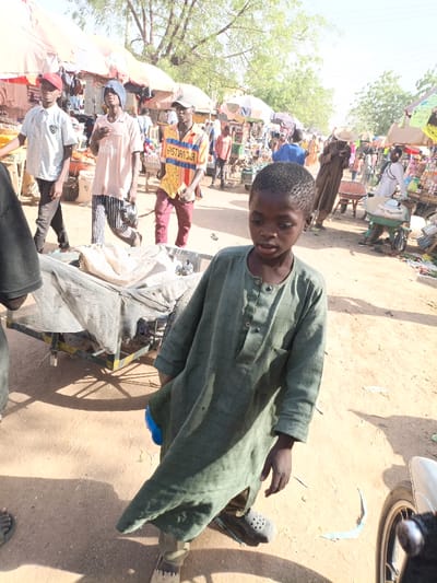 This image depicts a bustling outdoor market scene in Kawaji, Nigeria, under bright daylight.
In the immediate foreground on the right, a young boy with dark skin and short dark hair is standing, looking slightly to his left. He wears a loose-fitting, long-sleeved dark green tunic with buttons and dark sandals. A blue object is partially visible, clutched in his left hand near his hip. To the left of the boy, a portion of a person's foot in sandals and a dark-sleeved arm are visible. A two-wheeled handcart, covered with a light-colored fabric or netting, is situated behind the boy in the mid-ground.
Behind the foreground subjects, a dirt path extends into the background, flanked by numerous market stalls. Three young men are walking from left to right across the mid-ground. The first wears a light polo shirt, dark pants, and a red baseball cap. The second wears a light pink t-shirt, striped pants, and a blue head covering, carrying a dark bag. The third wears a yellow and red patterned t-shirt with "CHRISTIAN DIOR" text, reddish-brown pants, and a light-colored baseball cap.
The background features a dense collection of market stalls with various light-colored awnings, displaying goods that are indistinguishable at this distance. Numerous other individuals are present, walking, standing, and interacting within the market. One person in the distant mid-ground wears a wide-brimmed hat and a long brown garment. Another person further back is pushing a green wheelbarrow laden with items. The ground is unpaved dirt, casting distinct shadows from the bright sunlight. Sparse trees are visible above the market stalls in the far background. A partial view of a white motorcycle wheel is visible in the bottom right corner of the frame.