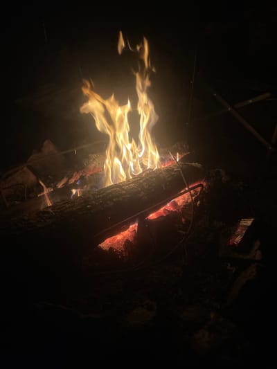 A campfire burns at night, illuminating logs and embers. The flames are bright yellow and orange, reaching upwards into the darkness. Some smoke is visible rising from the fire. In the lower right portion of the image, a small red object, possibly a package of matches, lies on the ground near the fire. The scene is dark, with the fire being the primary light source. The location is specified as Mala Kokhnivka, Ukraine.