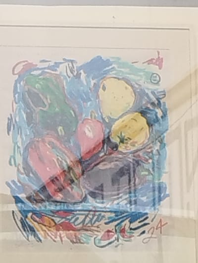 A framed, abstract still life artwork is depicted, featuring multiple fruits or vegetables rendered with loose, colorful brushstrokes. The artwork is predominantly blue in the background, with the objects appearing in shades of green, yellow, pink, red, and dark grey/purple. Specific forms include a large green elongated object on the top left, two round yellow objects on the top and middle right, a pinkish-red round object in the center, a larger reddish-pink elongated object on the bottom left, and a dark grey mass at the bottom center. The lower right corner of the artwork contains red markings, including the numbers "24". The artwork is enclosed within a simple, light-colored frame. Significant glare and reflections are visible across the glass covering the artwork, obscuring portions of the image and indicating the presence of ambient light sources in the room where it is displayed. The artwork is mounted on a light-colored wall. The specific location within Montevideo, Uruguay, where this image was captured is not visually discernible from the provided data, but it is presented as an indoor setting. No individuals or direct interactions are visible.
