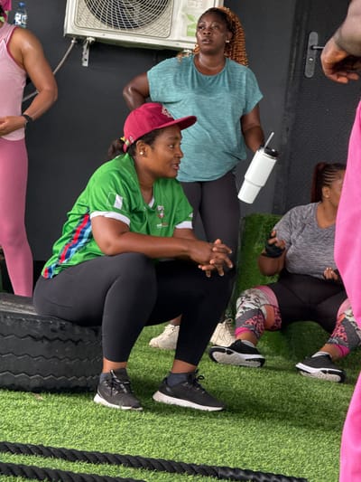 This image depicts an indoor fitness setting, possibly a gym, with artificial turf flooring. In the foreground, a Black woman wearing a green jersey with red and blue stripes on the sleeve, black leggings, and a red baseball cap is seated on a large black tire. Her hands are clasped in her lap. To her right, another Black woman in a grey t-shirt and black leggings with a colorful patterned design on the lower legs is also seated, her hands positioned in front of her. Behind them, a Black woman with curly brown hair, wearing a teal t-shirt, stands holding a white insulated tumbler. To the far left, the legs of a person wearing pink leggings are visible. In the background, a large air conditioning unit is mounted on the wall. The overall scene suggests a break or rest period during a workout session. The location is identified as Iddo Tudunwada, Nigeria.
