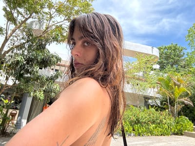 A woman with dark, wet hair and bangs stares to the left with a serious expression. Her skin tone is fair, and a portion of her shoulder is visible. Tattoos are visible on her back and arm. The background features lush green trees and a modern white building with rounded balconies. The sky above is partly cloudy. The overall lighting is bright and sunny.