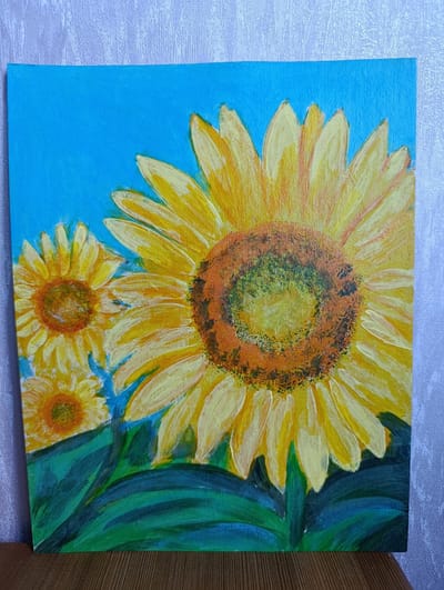 This is a painting of three sunflowers against a bright blue sky. The largest sunflower is in the foreground, its yellow petals radiating outwards from a textured brown and green center. Two smaller sunflowers are visible behind the main one, also depicted with yellow petals and darker centers. Lush green leaves fill the bottom portion of the painting, providing a grounding contrast to the vibrant yellow of the flowers and the vivid blue background. The painting appears to be an acrylic or oil on canvas, with visible brushstrokes that give it a textured, almost impasto quality. The lighting suggests a bright, sunny day, typical of late summer or early autumn. The style is expressive and somewhat impressionistic, focusing on color and form rather than precise detail. There is no visible text in the image. The setting is likely an outdoor scene, though the background's uniform blue could also suggest a studio setting. The overall mood is cheerful and vibrant.
