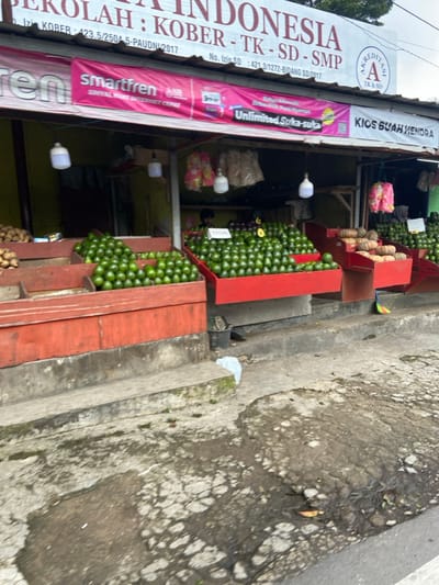 The image shows a fruit stand located in Lembang, Indonesia, at the coordinates -6.818 latitude and 107.613 longitude. The stand is fronted by a cracked, gray concrete sidewalk. The stand is covered by a wooden awning displaying signs in the Indonesian language. The sign on the right states “KIOS BUAH HENDRA.” The sign on the left reads “SEKOLAH INDONESIA KOBBER - TK - SD - SMP” and displays a pink and white “smartfren” logo. The stand is selling green avocados stacked in wooden crates with red borders. Several bags of what appears to be onions are visible inside the stand. There are two white, string lights hanging from the awning above the stand. A person appears to be working inside the stand, but their face is obscured by the counter. A broom is laying on the sidewalk in front of the stand.