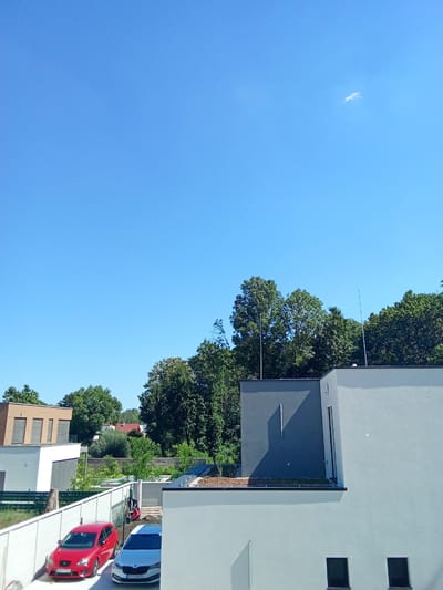 This image depicts a clear, bright day in Bratislava, Slovakia. The foreground features a white, modern-style building with dark-framed windows. To the left of this building, two cars are parked in a driveway enclosed by a white fence: a red hatchback and a white hatchback with a license plate that reads "ST 088GK". Behind the fence, another building with a brown facade and horizontal window shutters is visible.

The middle ground is dominated by lush green trees, forming a dense foliage backdrop. Above the trees, parts of other residential buildings with red roofs can be seen. Two tall, thin metallic poles, possibly lightning rods, extend upwards from the rooftops. The sky is a vibrant, cloudless blue, with a single wispy white cloud visible high in the upper right quadrant. The bright sunlight and clear sky suggest it is midday. The overall scene conveys a peaceful, residential setting on a sunny day.