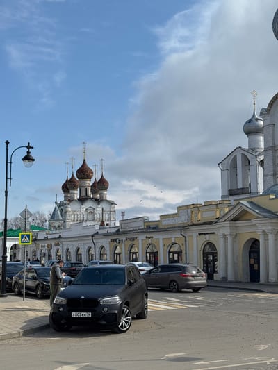 Captured on a sunny day in Novo-Nikol'skoe, Russia, the image showcases a street scene featuring several parked cars along a building with ornate architecture. The architectural style suggests a blend of classical and traditional Russian design, with arches, decorative facades, and domed roofs in the background. In the foreground, there are vehicles parked on the roadside. One individual is interacting with the vehicles; possibly parking, or talking on the phone. A pedestrian crossing is visible on the road. Street furniture, such as a lamppost, is present. The sky is partially covered with clouds, allowing ample sunlight to illuminate the scene.