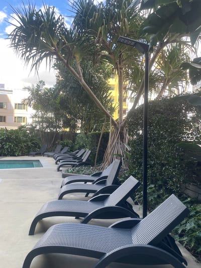 This image depicts an outdoor swimming pool area with multiple black plastic lounge chairs arranged on a concrete patio. Lush green foliage and palm trees surround the area, with a modern yellow building visible in the background. A tall, dark lamppost stands prominently in the foreground. The sky above is partly cloudy. The scene appears to be a tranquil setting within a residential or hotel complex, likely in a warm climate. Given the presence of such tropical vegetation and modern architecture, this scene could be located in a city like Brisbane, Australia, known for its outdoor lifestyle and subtropical climate.