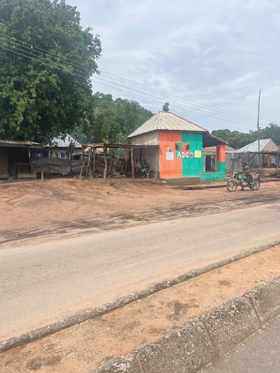 The image depicts a roadside scene in Minda, Nigeria. A small, colorful building, painted in orange and green, is prominently featured on the left side of the frame. The building has signage that appears to read "ADCA" and displays images that look like film reels. A corrugated metal roof extends over the structure and a shaded, open-air area in front, supported by wooden posts.
In front of the building, a motorcycle with two people on it is parked. Beneath the shaded area, several people are gathered, engaged in conversation or activity, though their faces are not clearly discernible. Further down the road, other simple structures made of corrugated metal and wood are visible, suggesting a semi-urban or rural setting.
The road itself is paved and appears to be a main thoroughfare, with a dirt shoulder bordered by a concrete curb. Lush green trees and vegetation line the background, indicating a tropical or sub-tropical environment. The sky is overcast with clouds, suggesting a diffused light, possibly indicating it is daytime, though the exact time of day is difficult to determine. The overall impression is one of everyday life in a Nigerian town.