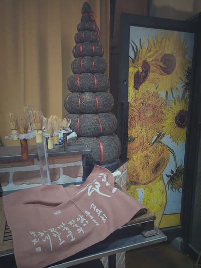 This image depicts a display featuring a towering stack of round, dark-colored pressed tea cakes, each tied with a red ribbon, arranged in a pyramid shape. To the left, a collection of traditional bamboo matcha whisks, known as "chasen," are displayed on stands. A small, patterned ceramic water pitcher is also visible. In the foreground, a folded, light reddish-brown cloth with white calligraphic writing is draped. Behind the tea stack, a framed print of Vincent van Gogh's "Sunflowers" painting is prominently displayed. The background consists of a golden-brown curtain. The setting appears to be an indoor retail space, possibly a tea shop or a cultural artifact store. There is no information in the image to pinpoint a specific location in Busan, South Korea.
