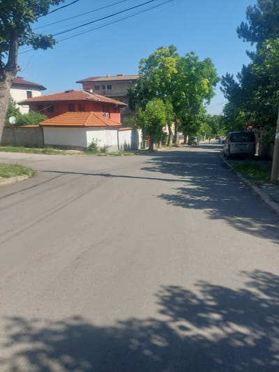 The image depicts a street scene in the city of Markovo, Bulgaria, under a clear blue sky. The road, paved with asphalt, is lined with trees casting shadows on the surface. On the left side of the street, residential buildings with tiled roofs are visible, separated from the road by a low wall and some vegetation. One building features a prominent orange-tiled roof. On the right side of the street, a silver van with license plate PB 9317 AB is parked. Further down the street, several other vehicles are present, and one person can be seen cycling. The overall environment suggests a quiet, residential area on a sunny day.