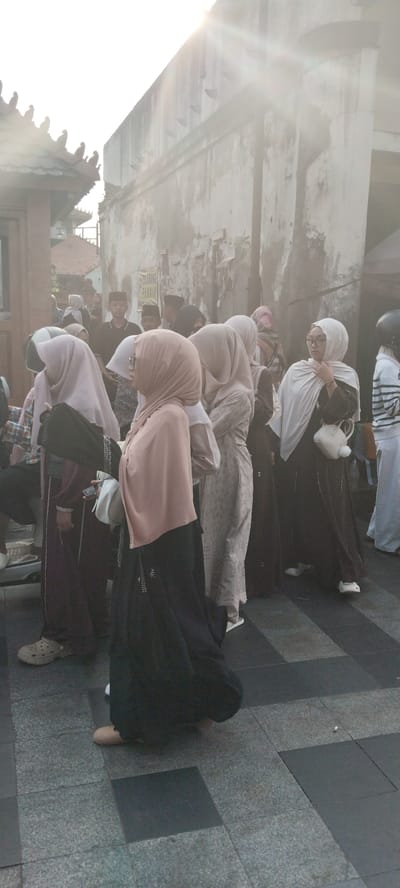 This is a street-level photograph taken in the city of Kauman, Indonesia, during daylight hours, possibly in the late afternoon due to the strong backlight and lens flare. The scene depicts a gathering of people, many of whom are wearing hijabs and modest clothing, suggesting a culturally conservative environment. The architecture visible includes traditional Indonesian tiled roofs and a weathered, textured wall. The ground is paved with patterned tiles. The overall impression is that of a public space where people are socializing or waiting.