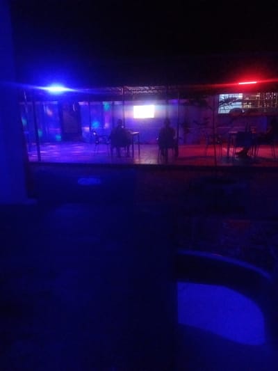 The image depicts an outdoor seating area at night, illuminated by blue and red neon lights. In the background, a covered structure with glass walls reveals a room with tables and chairs, where two individuals are seated. One person is visible from the back, facing a television screen. The environment appears to be a dimly lit establishment, likely a bar or restaurant, in Sabon Gari, Nassarawa, Nigeria. The foreground is dominated by a dark, unlit area with a reflective blue surface, possibly water or a wet floor.