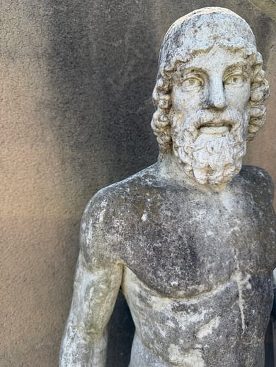 The image shows a weathered stone statue of a male figure, possibly depicting a historical or mythical character. The statue is laying on a textured stone surface. The statue has a detailed head with curly hair and beard. The body is partially visible, showing an arm and torso. The statue is located outdoors, presumably in Oullins-Pierre-Bénite, France.