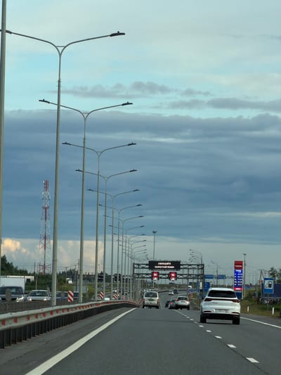 The image depicts a highway scene in Skotnoe, Russia, captured during daylight hours. The sky is overcast with dramatic, streaky clouds, suggesting potentially unsettled weather. The perspective is from within a vehicle, looking forward along the right lane of a multi-lane highway.
The main subjects are the vehicles on the road and the surrounding infrastructure. A white SUV is prominent in the foreground, followed by other cars and trucks. The highway is flanked by numerous streetlights mounted on tall, curved poles, creating a repeating pattern. A metal barrier with red and white reflective markers separates the lanes from the shoulder. In the distance, gas station signs for "TEBOIL" are visible, indicating commercial activity alongside the highway. One sign displays fuel prices: 65.00, 69.73, 72.64, 79.37, and 79.57, likely in rubles per liter.
A digital traffic sign displays "СОБЛЮДАЙТЕ СКОРОСТНОЙ РЕЖИМ" (Observe the speed limit) along with a "90" indicating the speed limit in kilometers per hour. This suggests an active traffic control system is in place. Further down the road, more cars are visible, suggesting moderate traffic. A red and white radio tower can be seen in the middle ground, adding to the industrial or infrastructure elements of the scene. The overall mood is functional and typical of a highway commute, with no strong emotional cues. The license plate of the white SUV is partially visible as T632XY 198.