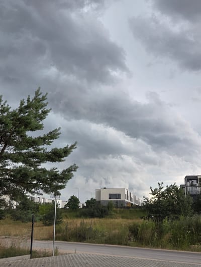 The image depicts an outdoor scene with a cloudy sky dominating the upper portion. In the lower part of the image, there are some trees, a road, and modern buildings with white facades and dark windows. The lighting suggests it might be late afternoon or evening, as the sky is overcast and the light is diffused. The weather appears to be gloomy or on the verge of rain, with heavy grey clouds. The scene is set in an urban or suburban environment, likely a residential area given the architecture of the buildings. There are no people visible in the image. The overall mood is somewhat somber due to the weather conditions. The road is paved and runs alongside a grassy area with some vegetation and a fence.