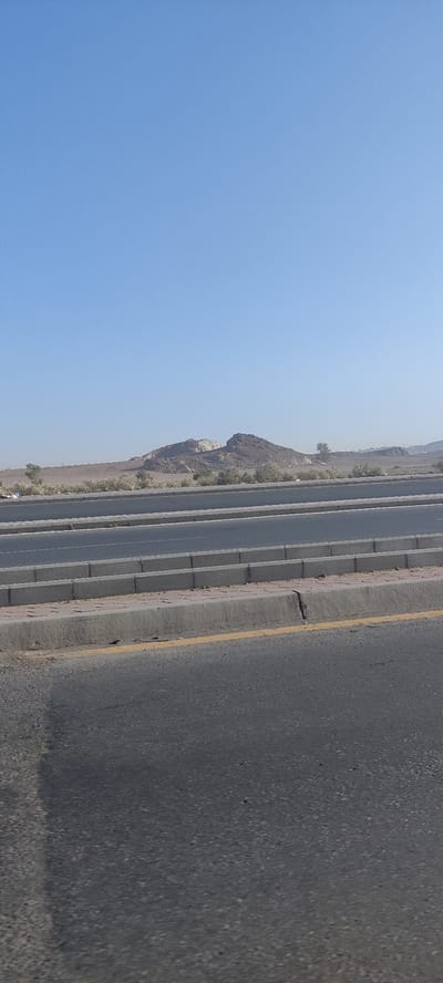 The image shows a view of Jabal Al-Nour, a mountain near Mecca, Saudi Arabia, seen from what appears to be a highway in Jeddah. The sky is clear and blue. The mountain is in the background, appearing brown with lighter-colored rock formations on its peaks. In the foreground, there is a multi-lane road with a concrete median and asphalt surfaces. The road takes up a significant portion of the lower half of the image. A yellow line marks the edge of the road. The lighting suggests it is daytime. There are few shrubs and trees in the desert landscape between the mountain and the road. No people are visible.