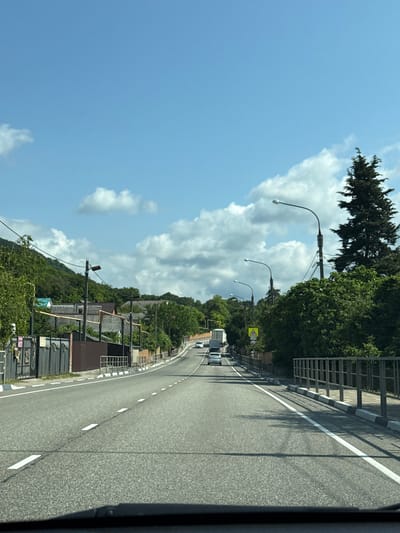 The image depicts a view from a vehicle traveling on a paved road that curves to the right. The road has white lane markings and a metal guardrail on the left side. Beyond the guardrail, there are trees and some buildings with tiled roofs. On the right side of the road, there's a steep, forested hill. Several streetlights with curved arms are visible along the road. The sky is clear blue with scattered white clouds. The scene appears to be a sunny day, possibly late morning or early afternoon, given the brightness and shadows. The location is Olginka, Russia. There is a yellow triangular road sign with a black symbol on the left side of the road. No people are clearly visible in the image.