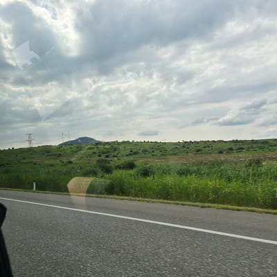 The image depicts a landscape scene with a road in the foreground and a grassy hill with vegetation and power lines in the background. The sky is filled with dramatic clouds, suggesting it might be a partly cloudy day. In the distance, a hill is visible, with some structures that appear to be power transmission towers. The road is asphalt with a white line marking the lane. There are also some hay bales visible on the side of the road. The scene is likely captured from a moving vehicle due to the slight blur and the angle of the shot. The image does not contain any people or specific landmarks that would allow for definitive identification of the location within Zheleznovodsk, Russia, beyond it being a rural or roadside landscape.