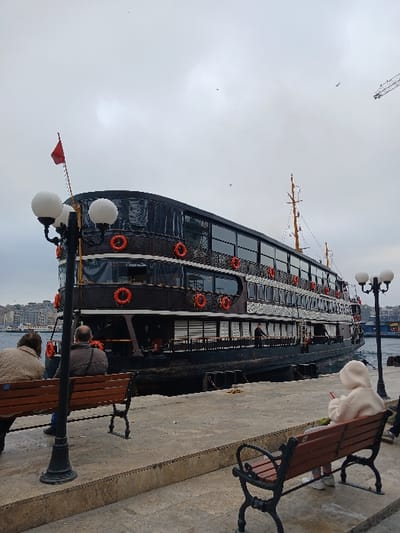 The scene takes place on a pier in İstanbul, Türkiye, near the coast, at the coordinates 41.022° N, 28.976° E. A black ferry boat is docked at the pier. There are three people in the scene: two sitting on a bench by a lamppost in the foreground and one seated on a bench in the lower right corner of the image. The two figures in the foreground are facing each other, and the third figure is facing the ferry boat. The scene is overcast and the sky is gray. The ferry boat is multi-level and has red life preservers around the railings. There is a small red flag flying from a pole at the top of the pier.
