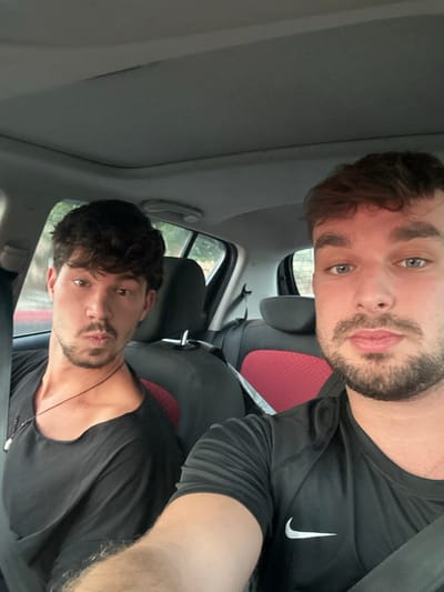 The image depicts two adult males seated inside a vehicle. The male in the foreground, positioned on the right, has short brown hair, light blue eyes, and a trimmed beard; he is wearing a black Nike athletic shirt with a white swoosh logo. He is secured by a seatbelt and directly faces the camera. The male in the background, positioned on the left, has dark curly hair, brown eyes, and stubble; he is wearing a black, wide-neck shirt and a thin dark necklace with a small pendant. His lips are pursed, and he is also secured by a seatbelt. The vehicle's interior features a light grey headliner and dark-colored seats, with a visible red accent panel on the backrest of the front passenger seat. The exterior environment, visible through the blurred rear window, shows indistinct green foliage. No specific landmarks are discernible to identify a precise location within the city of Rishon LeZion, Israel.