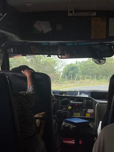 The image captures an interior perspective from a vehicle, likely a bus, looking forward through the windshield. A passenger, wearing a black and white striped shirt, is seated in the foreground, viewed from behind, with their right hand gripping the top of the seat in front of them. A portion of another passenger's shoulder is visible on the far right.

Above the windshield, the dark ceiling features a centrally located rearview mirror. To the right of the mirror, a black circular fan is mounted. On the driver's side, a white rectangular sign reads, "BUS MUST NOT STAND FORWARD OF WHITE LINE WHILE BUS IS IN MOTION." Adjacent to it is a yellowish-brown rectangular sign, and two white papers are affixed to the ceiling.

The dark-colored dashboard below the windshield is equipped with various controls, gauges, buttons, a radio unit, and air vents. A yellow diamond-shaped sticker and other warning labels are present. A blue rectangular box sits on top of some fabric or clothing on the passenger side of the dashboard, with a red object labeled "Electro" visible beneath it.

Through the front windshield, a paved road, marked by a white line on its left edge, is visible, curving gently to the left. The road is flanked by dense green foliage and trees, indicating a natural environment. A fence constructed from rustic wooden posts runs parallel to the road on the left side.

This scene is consistent with travel through a rural or semi-rural area within El Congo, El Salvador, characterized by its abundant vegetation and local transportation.
