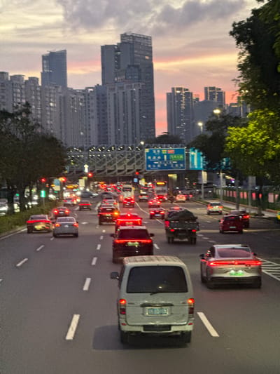 The image shows a traffic jam on a multi-lane highway in the Nanshan District of Shenzhen, China, at dusk. Skyscrapers are visible in the background with a colorful sunset sky above. Cars and trucks are densely packed in the lanes with their tail lights illuminated. A green highway sign is visible among the vehicles. A bridge structure is visible on the left side of the road. Trees line parts of the roadway. The photo is taken from an elevated position, likely an overpass or building.