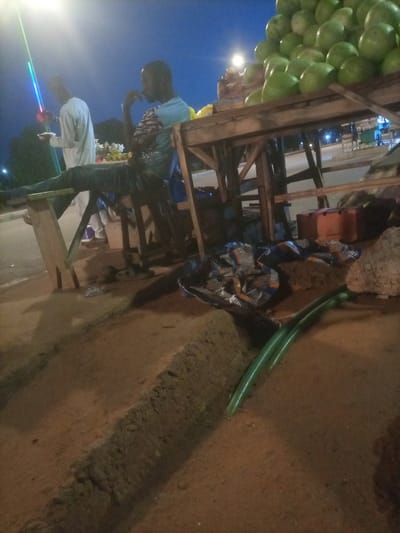 The image depicts an outdoor scene at dusk, likely in Wuro Dole, Nigeria. A vendor is partially visible, seated on a wooden bench with his legs extended, appearing to be resting. He is wearing a blue and white patterned shirt and dark pants. Beside him, another person in a white garment is standing, holding what appears to be food on a plate. In the background, a tall structure with colorful lights, possibly a street lamp or decoration, illuminates the scene. A large pile of green fruits, likely watermelons or similar produce, is stacked on a wooden table. The ground is dirt, with a shallow trench running through it. Various objects, including fabric, containers, and what looks like a green hose, are scattered around the area. The sky is a deep blue, indicating the transition from day to night.
