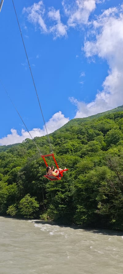 This vertical shot captures a thrilling outdoor activity in a scenic mountainous landscape. The central subject is a person, likely a woman, seated in a vibrant red swing suspended high above a river. She is wearing a yellow top and light-colored bottoms, and is holding a mobile phone in her hands, possibly recording or taking a picture. Her posture appears relaxed as she hangs suspended by multiple thick metal cables.

The setting is characterized by a dramatic natural environment. In the background, a steep, densely forested mountain rises under a bright blue sky adorned with scattered white clouds. The trees on the mountainside are lush and green, indicating a warm, possibly summer, season. Below the swing, a wide river flows, its water appearing a turbid light brown or greyish-white, suggesting a strong current or glacial melt. The riverbank on the far side is also heavily forested.

The image conveys a sense of adventure and exhilaration, with the person enjoying the panoramic views from a considerable height. The bright daylight and clear sky suggest excellent weather conditions for such an activity. The scene is set in a picturesque natural area, specifically Bzypta, Abkhazia. No visible text is present in the image.
