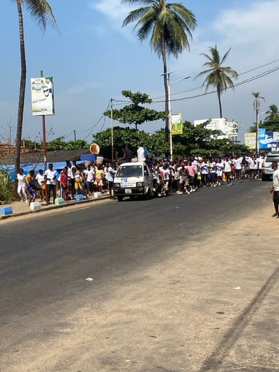 This media file captures a vibrant street scene in Freetown, Sierra Leone, on a bright, sunny day. The image looks down an asphalt road, with a large crowd of people gathered along its left side. Many individuals in the crowd are wearing white shirts, suggesting a unified group or a specific event. The gathering extends into the distance, indicating a substantial assembly, possibly a parade or a public demonstration.

In the mid-ground, a white utility vehicle or small truck is parked among the crowd, with people standing on its roof and alongside it, participating in or observing the event. One person near the truck is holding a large, light-colored, round object, resembling a basket or a hat.

The environment is distinctly tropical, featuring numerous tall palm trees and lush green foliage lining the street. Utility poles with overhead power lines are also visible. In the background, buildings can be seen beyond the tree line. The sky is clear blue with minimal clouds.

Several street signs and billboards are visible. A prominent green and white sign clearly displays the text "BAZZ MONEY." Another sign further down the street appears to show partial text, possibly "PREPAR[ATION] CAR[ES]". The road surface itself is mostly clear of other vehicles, indicating the street has been taken over by the crowd for the event. The atmosphere appears lively, consistent with a public gathering.