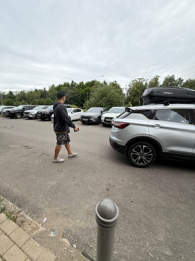 In Moskovskiy, Russia, a man wearing a black baseball cap, a dark hooded sweatshirt, and camouflage shorts walks across a paved parking lot. He is holding a mobile phone in his right hand and looking to his right. In the background, several cars are parked in a row, including a silver SUV with a black roof cargo box, a black SUV, a white SUV, and other vehicles. Trees and buildings are visible in the distance, under an overcast sky.