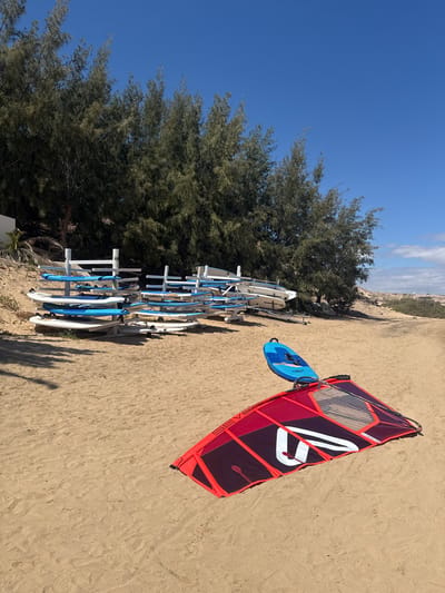 The image captures a bright, sunny scene on a sandy beach, likely in Pájara, Spain. In the foreground, a vibrant red and dark purple windsurfing sail lies on the sand, angled towards the left. It features a prominent white, stylized logo that resembles a '7' or 'V' with a line, and smaller text "SEVEN..." is partially visible along its base. Next to the sail, a blue windsurfing board with white accents also rests on the sand.
To the left, a long rack holds numerous white and blue surfboards or smaller windsurfing boards, indicating a rental or storage facility for water sports equipment. Behind the rack, a dense line of tall, dark green pine-like trees stretches across the middle ground, providing a natural backdrop.
The background on the right reveals a distant sandy landscape with low vegetation and subtle hints of buildings or structures on a slight incline. The sky is a clear, bright blue with a few faint white clouds. Strong shadows cast on the sand suggest bright sunlight, indicating it is likely mid-day. The overall impression is one of leisure and outdoor recreation in a warm, coastal environment.