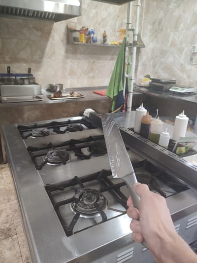 The image depicts a close-up view of a person's hand holding a large kitchen knife over a stainless steel commercial gas stove. The stove has four burners, with the knife's blade positioned above the front left burner. The kitchen environment appears to be a commercial setting, possibly a restaurant or catering establishment, with stainless steel surfaces and professional kitchen equipment visible in the background. There are condiment bottles and other supplies on a shelf in the background. The lighting suggests an indoor setting, and the overall impression is one of food preparation. The image does not provide any specific visual cues that would allow for the identification of the city of Supsekh, Russia.