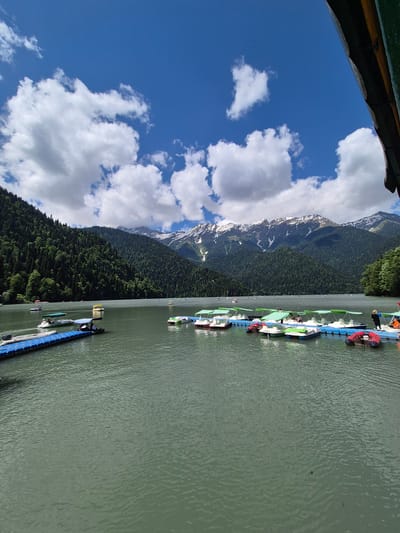 The image depicts a scenic view of Lake Ritsa in Abkhazia, a resort town known for its natural beauty. The lake is calm and reflects the surrounding mountains and sky. Several boats, including pedal boats and smaller motorized boats, are docked at a pier, indicating a recreational area. Lush green forests cover the slopes of the mountains that rise behind the lake, with snow-capped peaks visible in the distance. The sky is partly cloudy, with large white clouds scattered across a vibrant blue expanse. The overall impression is one of a serene and picturesque natural landscape, popular for outdoor activities.