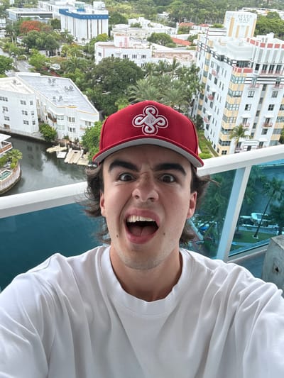 A person is taking a selfie on a balcony in Miami Beach, Florida, United States. The person is wearing a red baseball cap with a white clover-like design, and a white t-shirt. The person has dark hair and is making a funny face with their mouth open. The backdrop shows a waterway and buildings, and the “Helen Mar” building is on the left.