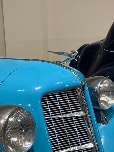 A close-up shot depicts the front of a vintage automobile. The vehicle has a light blue body, chrome grill, and prominent chrome headlights. An additional chrome ornament is visible on the side. The background is a plain, light beige wall. The scene appears to be indoors, possibly within a museum or private collection. The location is not visually discernible from the image.