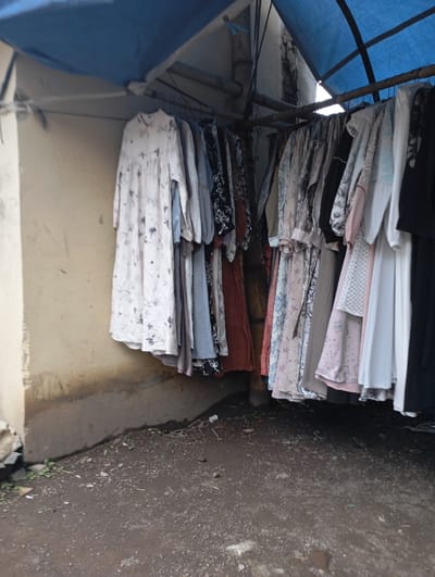The image shows a row of clothing hanging from a metal pole under a blue tarp. The clothing is mostly women's dresses and tops, and they are all in various patterns and colors. The pole is attached to a concrete wall and a pillar. The scene appears to be in a narrow outdoor space, likely an alley or a market stall, with dirt floor and a concrete curb. The scene is located in Kutawaringin, Indonesia, at the coordinates -6.974, 107.519.